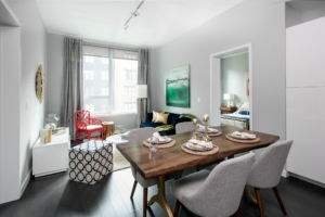 Apartment living room with 10' ceilings, large window for natural light, blue 3-seat sofa, adjacent a dining area set for four